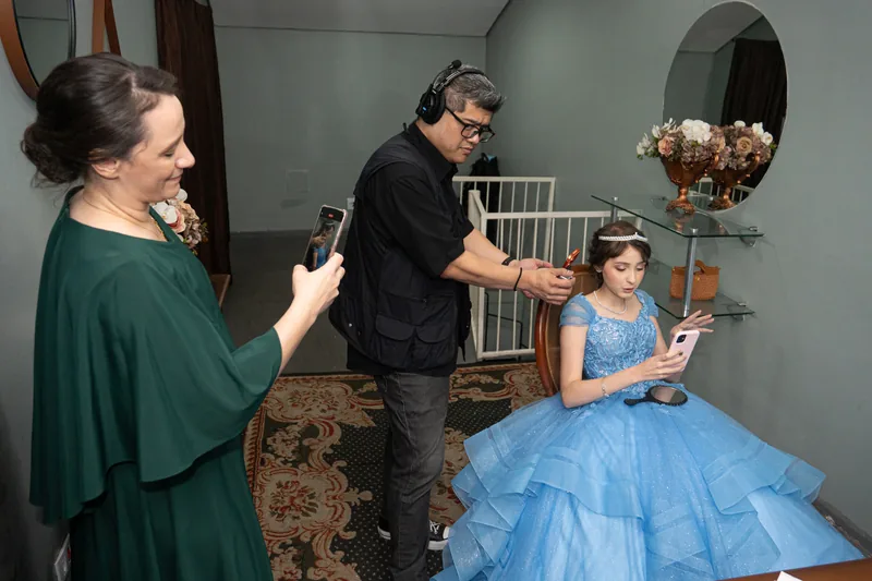 Camarim dos 15 anos: a debutante sentada de vestido azul enquanto a produção ajusta os últimos detalhes e a mãe filma pelo celular