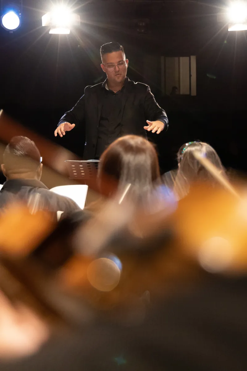 Maestro visto de trás, sob os refletores, diante da orquestra