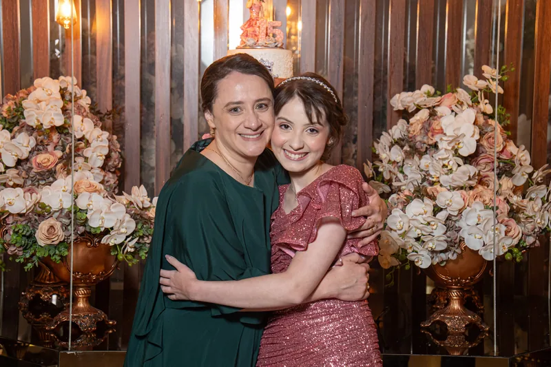 Mãe e filha maquiadas, abraçadas diante do bolo de 15 anos