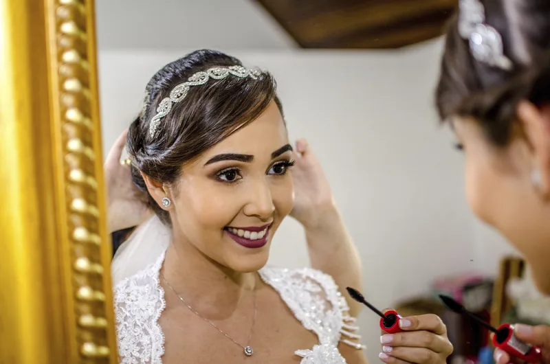Noiva sorrindo enquanto recebe os retoques finais diante do espelho iluminado